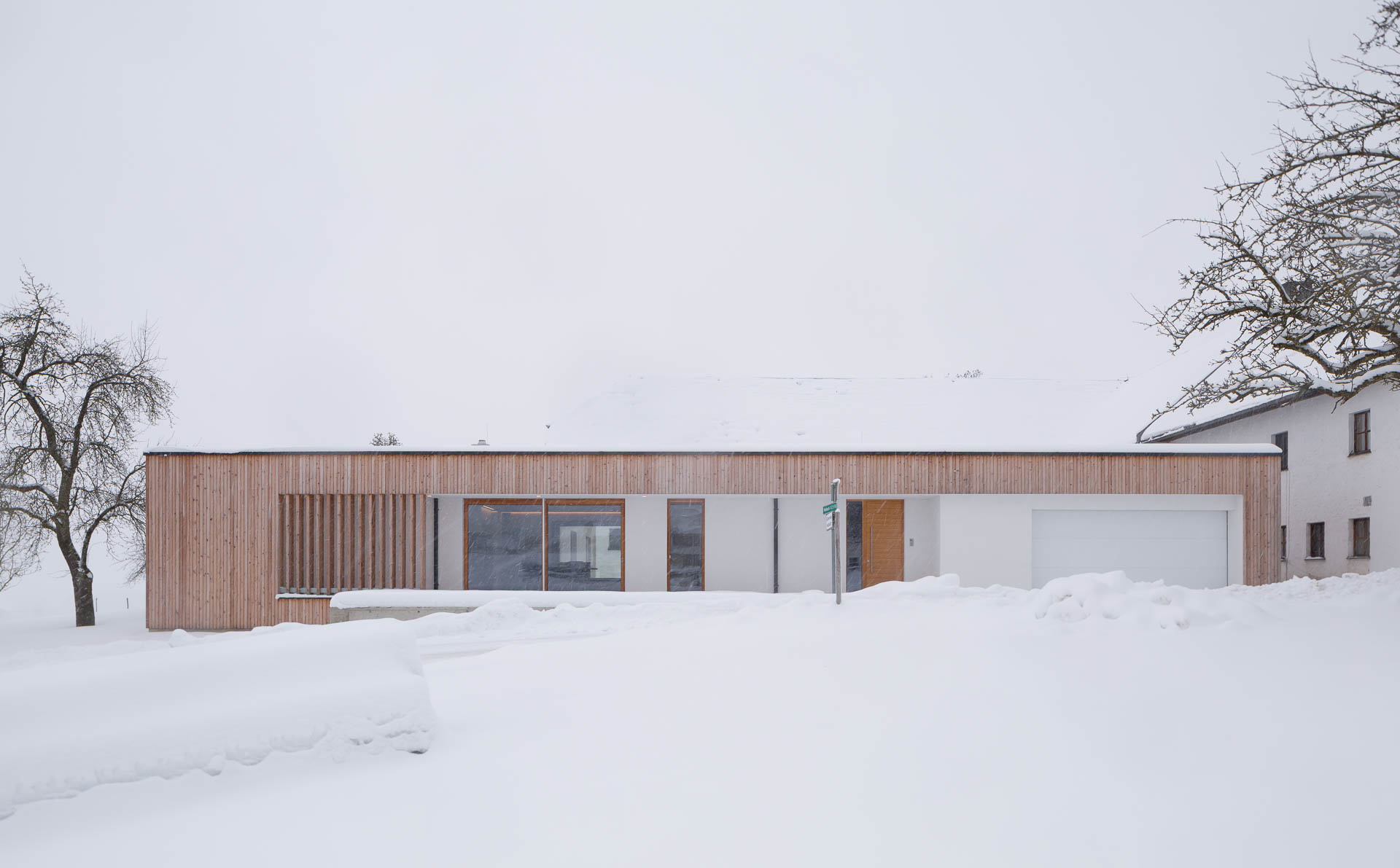 Bungalow Frontalansicht im Winter mit viel Schnee, dicke Schneedecke, warme Holzfassade als Kontrast, minimalistische Architektur, große Glasfenster, Holzlamellen,